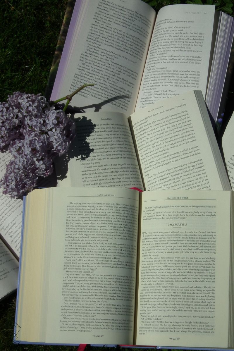 open books lying scattered on grass, with lilac flowers on left side.
