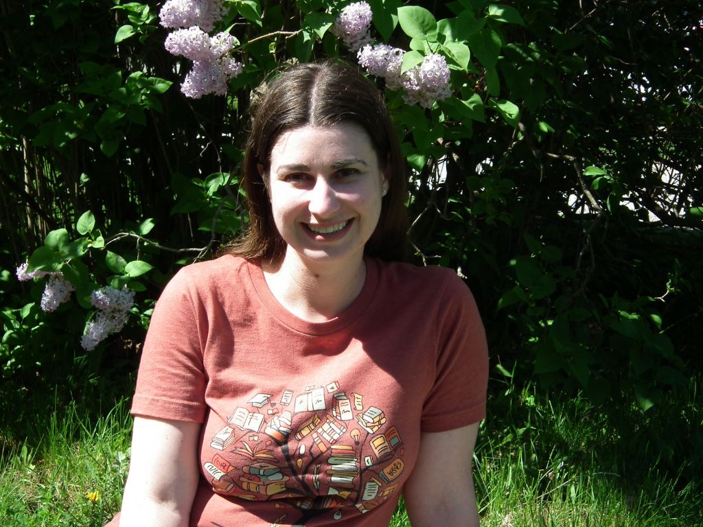 brunette woman smiling, wearing a red-orange bookish t-shirt, with lilac bush in background.
