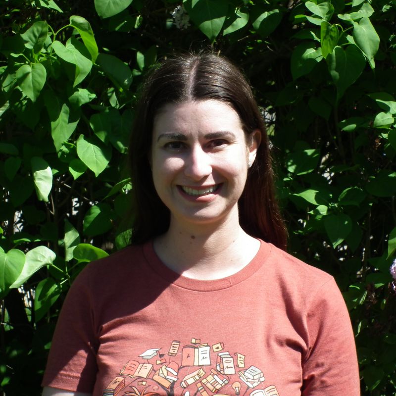 brunette woman in red-orange bookish t-shirt, with leaves in background.